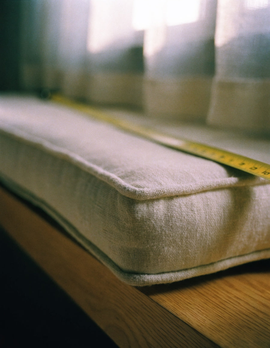 A piped boxed-edge linen bench cushion on an oak window seat, tape measure running along its back edge