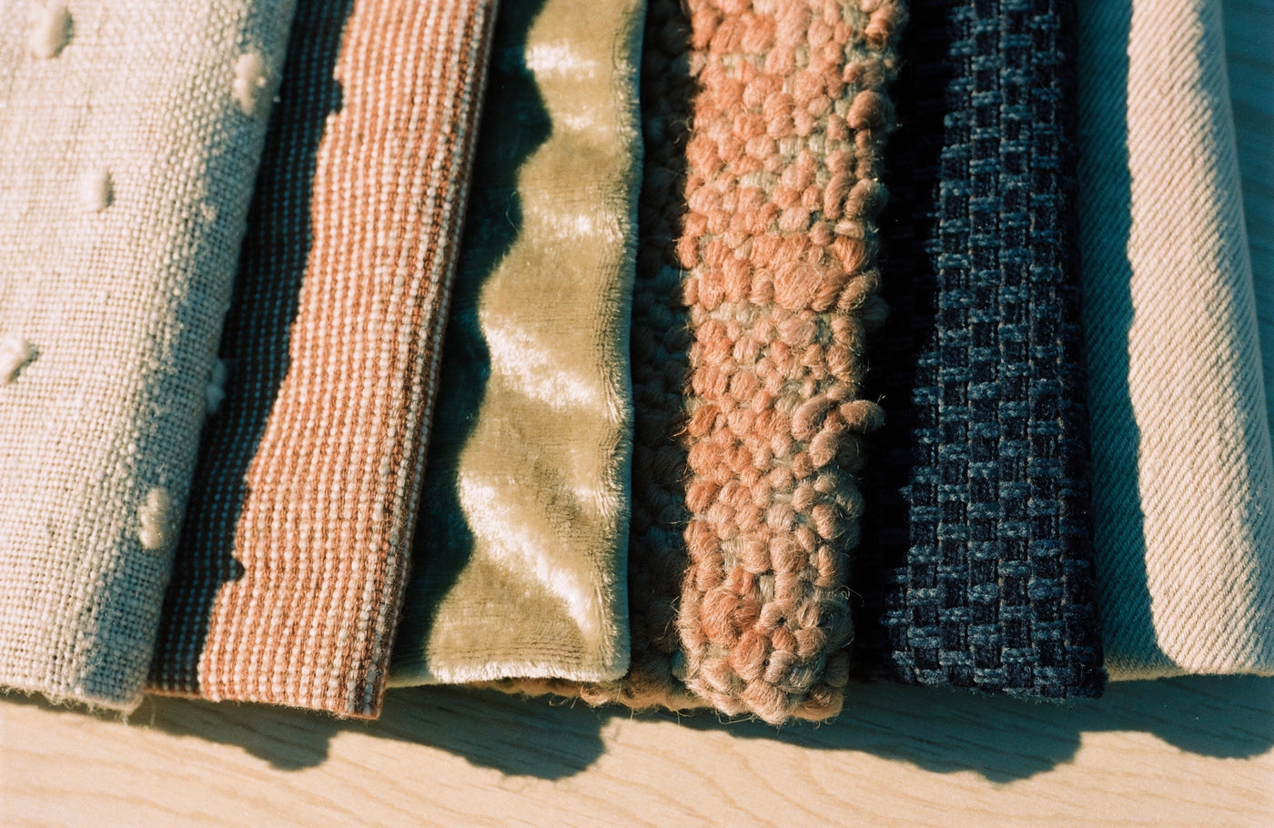 Six upholstery fabric swatches laid edge to edge, raking light revealing linen slubs, velvet pile and bouclé loops