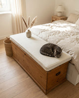 Cat sleeping on a cream cushion on a wooden chest at the foot of a bed